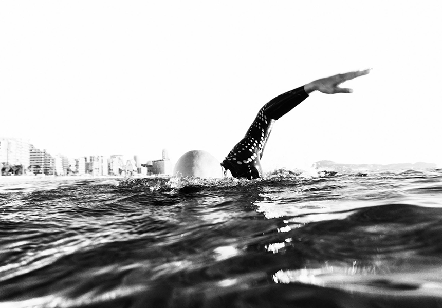 Foto em preto e branco de um homem nadando competitivamente no oceano em um terno molhado e óculos de proteção.
