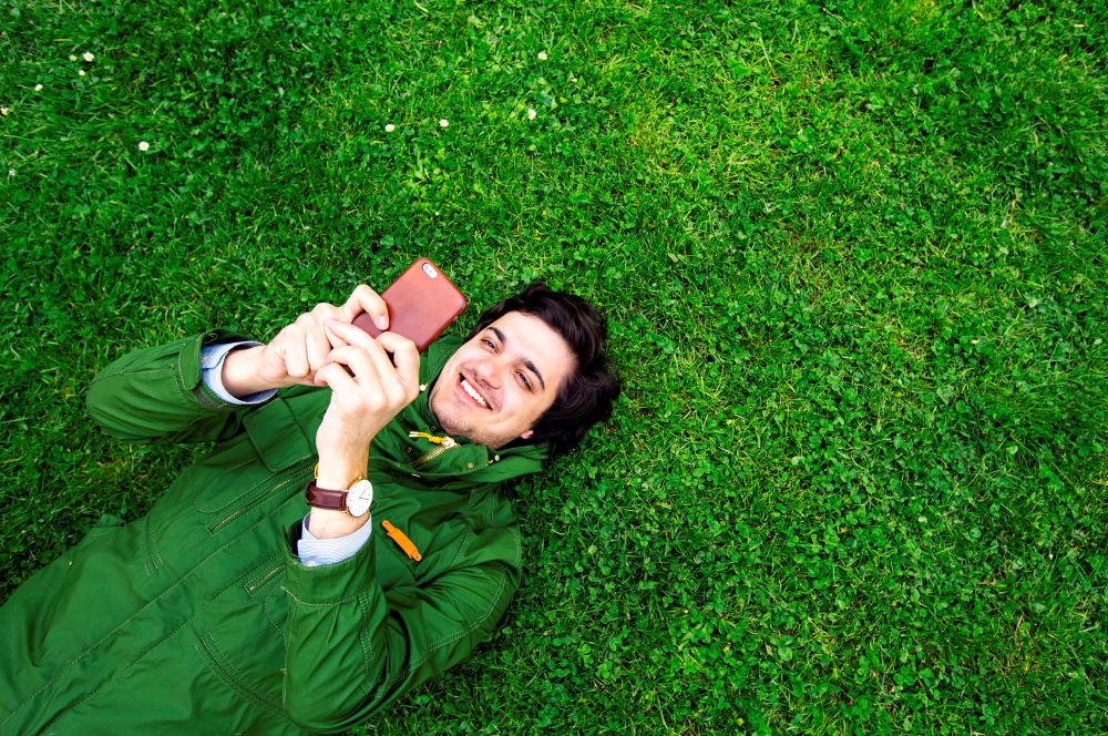 Man laying down on the grass, smiling at his phone.
