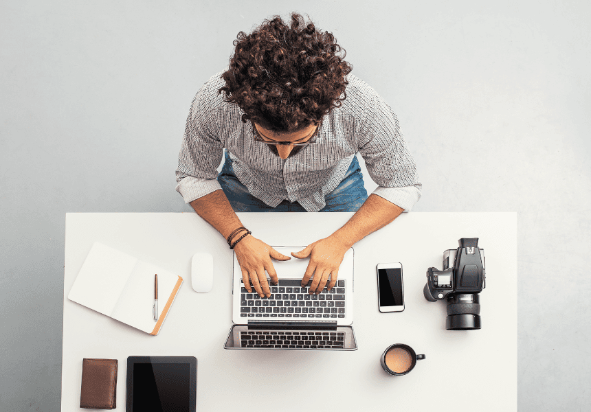 Persona trabajando en un escritorio vista desde arriba, usando un portátil. Sobre la mesa hay una libreta con un lápiz, un mouse, un teléfono móvil, una cámara, una taza de café y una tablet.