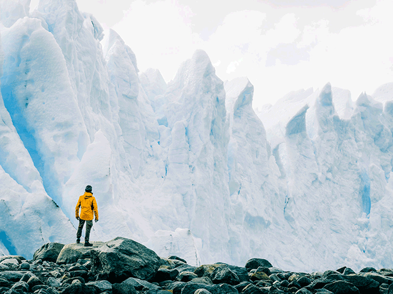 A hiker stands on rocks looking at a large iceberg