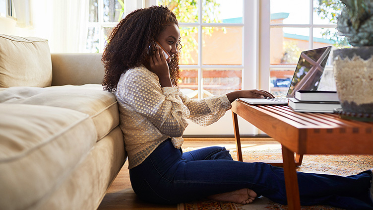 Young woman sitting on the floor taking a phone call whilst browsing on her laptop.