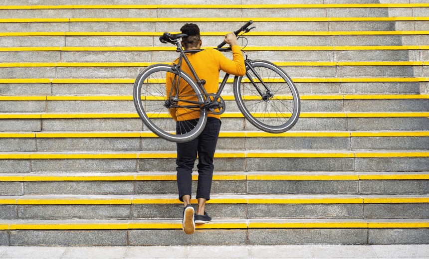 Young man walking up stairs carrying a bike on his back.