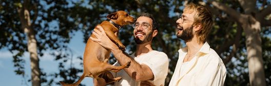 Two men laughing outdoors as one holds up a brown dog among trees.