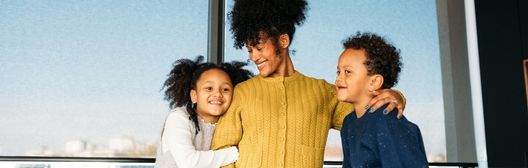 Mother standing with her arms around two smiling children in front of a large window.