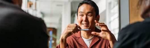 Smiling man in a red shirt holding a pen while speaking with two people in a bright office setting.