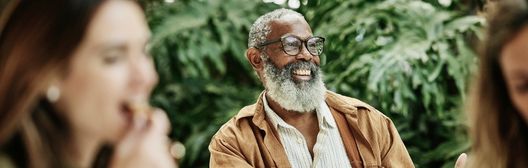 Smiling older man with glasses and a grey beard sitting outdoors with people blurred in the foreground.