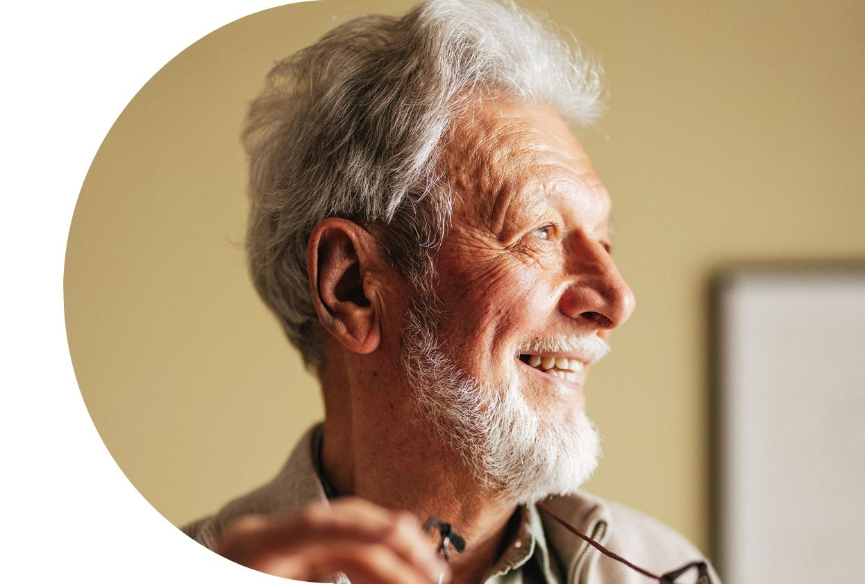 An older white-haired man smiles while looking off to the side and holding his glasses.