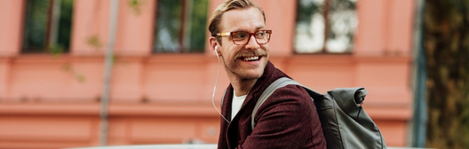 Smiling man with glasses and a backpack looking over his shoulder outdoors.