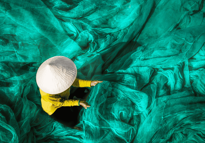 Person wearing a conical hat sits among layers of turquoise fishing net