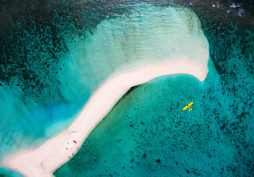 Aerial view of a white sandbar curving through clear turquoise water, with a small yellow aircraft visible nearby. 