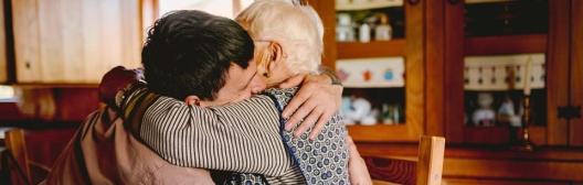 A man hugs an older woman seated at a kitchen table.