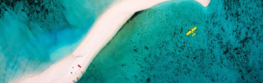 從空中俯瞰，一道白色沙洲蜿蜒穿過清澈的碧綠海水，附近可見一架黃色小型飛機。