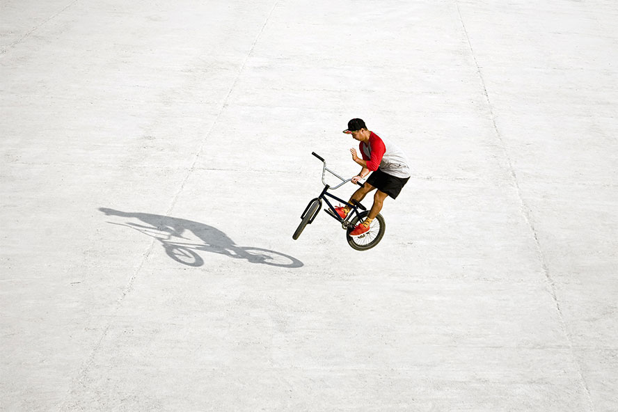 Against a white background, a man in baseball cap performs a trick on his bike while his shadow is prominent next to him.
