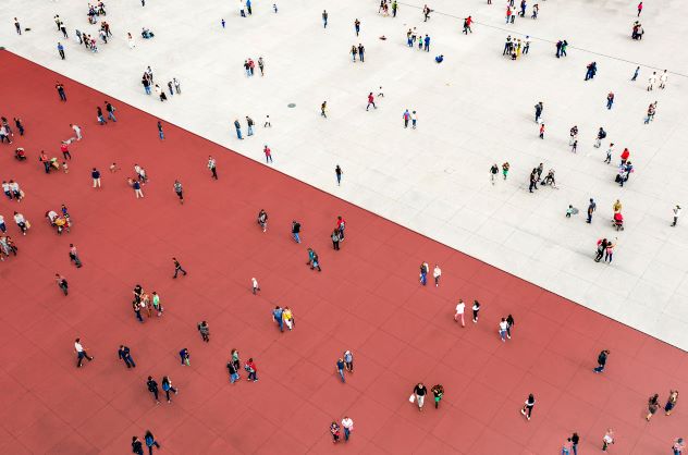 Aerial view of a large open plaza divided diagonally into two sections: one with red paving and the other with light grey paving. Numerous small groups and individuals are scattered across both areas, creating a sense of movement and activity.
