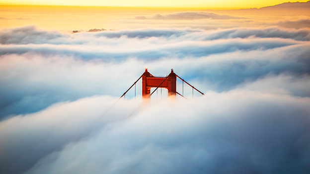 Top of a suspension bridge emerging through thick clouds at sunrise, with warm golden light in the background.