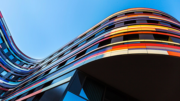 Low-angle view of a modern building with flowing, wave-like architecture featuring colorful horizontal panels against a clear blue sky