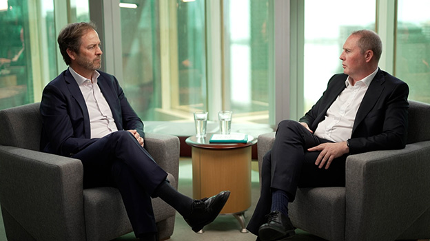 Two people in business attire seated across from each other in a modern office setting with large glass windows, a small table between them with two glasses of water.