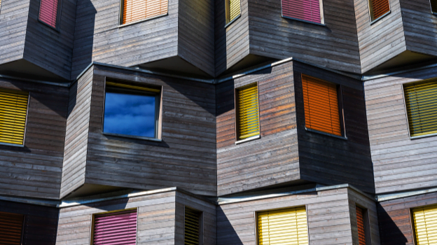 A modern building façade made of dark wooden panels with multiple protruding box‑shaped windows. Each window has brightly coloured blinds—yellow, orange, pink, and blue—creating a geometric, fragmented pattern.