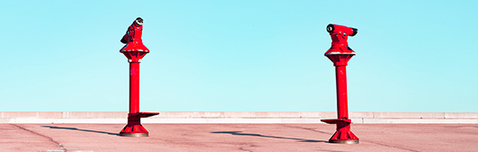 Wide image of two bright red coin‑operated viewing binoculars mounted on a paved waterfront promenade under a clear blue sky, with a bird perched on the left viewer.