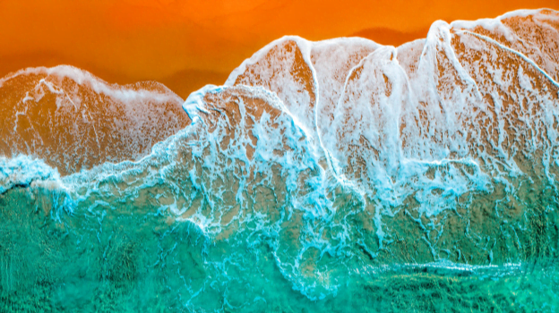 Aerial view of turquoise ocean waves crashing onto a bright orange shoreline, creating white foam patterns where the water meets the sand.