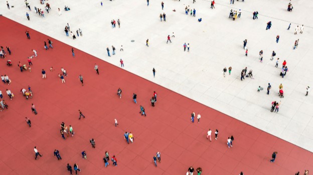 Aerial view of a large open plaza divided diagonally into two sections: one with red paving and the other with light grey paving. Numerous small groups and individuals are scattered across both areas, creating a sense of movement and activity.