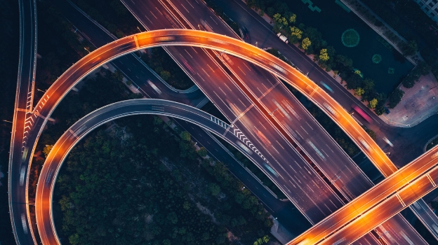 Aerial view of a multi-level highway interchange at night, with curved overpasses illuminated by orange light trails from moving traffic.