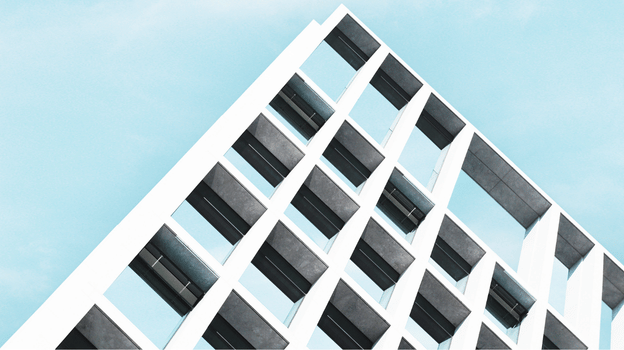 Photograph of a modern building facade featuring a grid of rectangular windows and white structural beams against a clear blue sky.