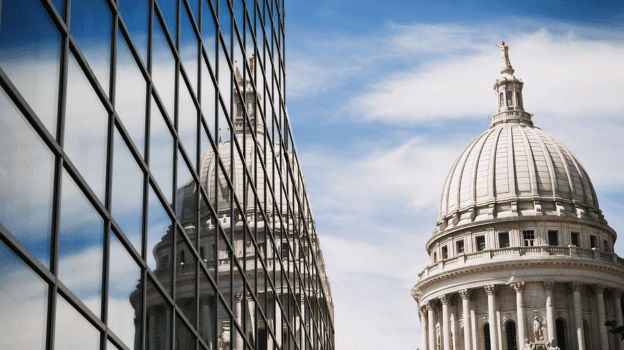 Historic domed capitol building reflected in the windows of a modern glass office tower under a blue sky.