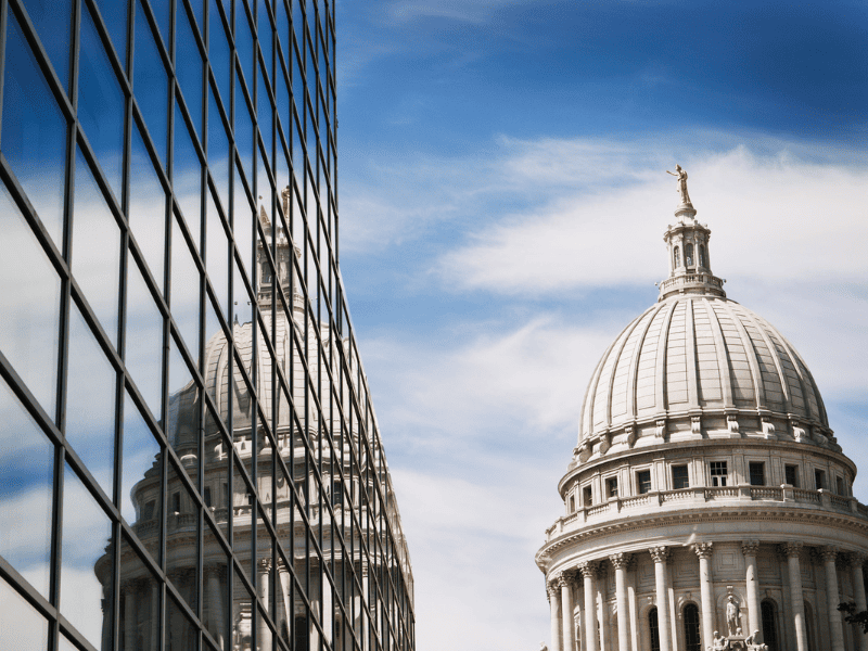 Historic domed capitol building reflected in the windows of a modern glass office tower under a blue sky.