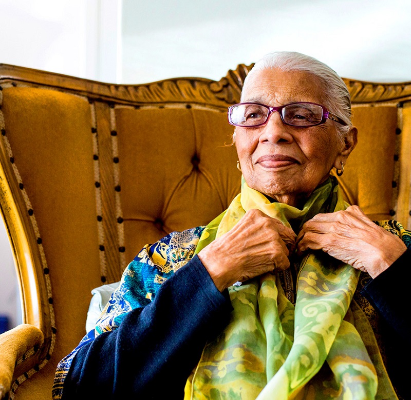 A latter middle aged woman adjust her silk scarf while sitting in a high back arm chair