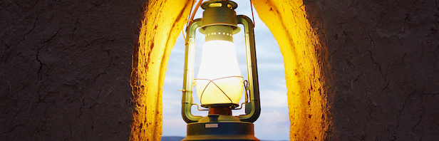 A person wearing a bright yellow jacket and winter hat stands on a rocky ground facing a massive wall of blue and white ice. The figure is small in comparison to the towering glacier, emphasizing the scale of the icy landscape.