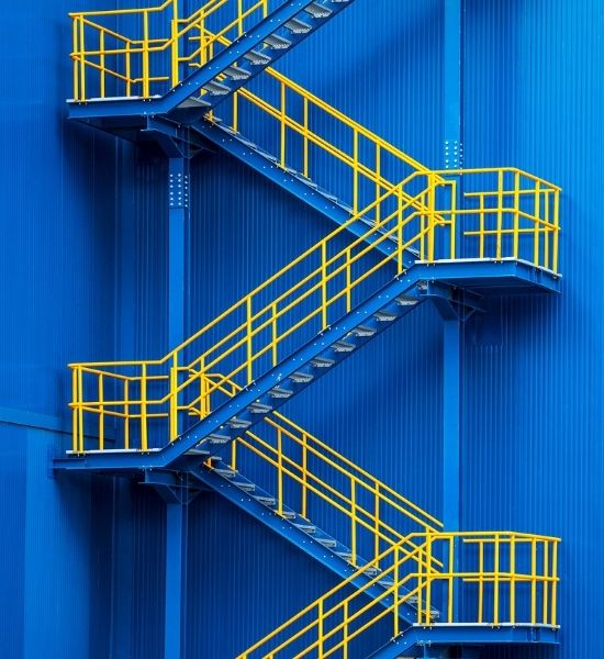 Zigzagging industrial metal staircase with yellow railings against a blue building exterior.