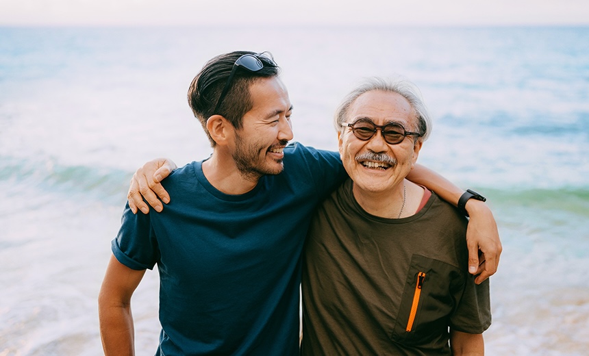 Two people standing together at the shoreline with their arms around each other, looking toward one another.