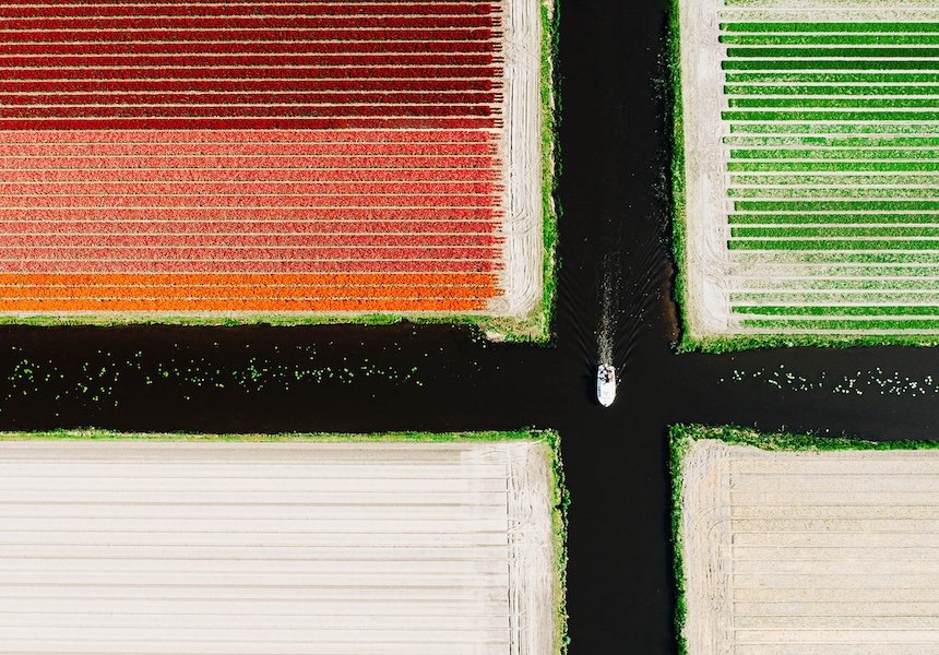 Aerial view of a patchwork of agricultural fields separated by intersecting dark waterways. A small boat travels through the center canal, creating a wake as it passes between the neatly striped fields