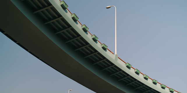 Two-lane elevated highway curving upward with streetlights along the edge; supported by large beams against a clear blue sky