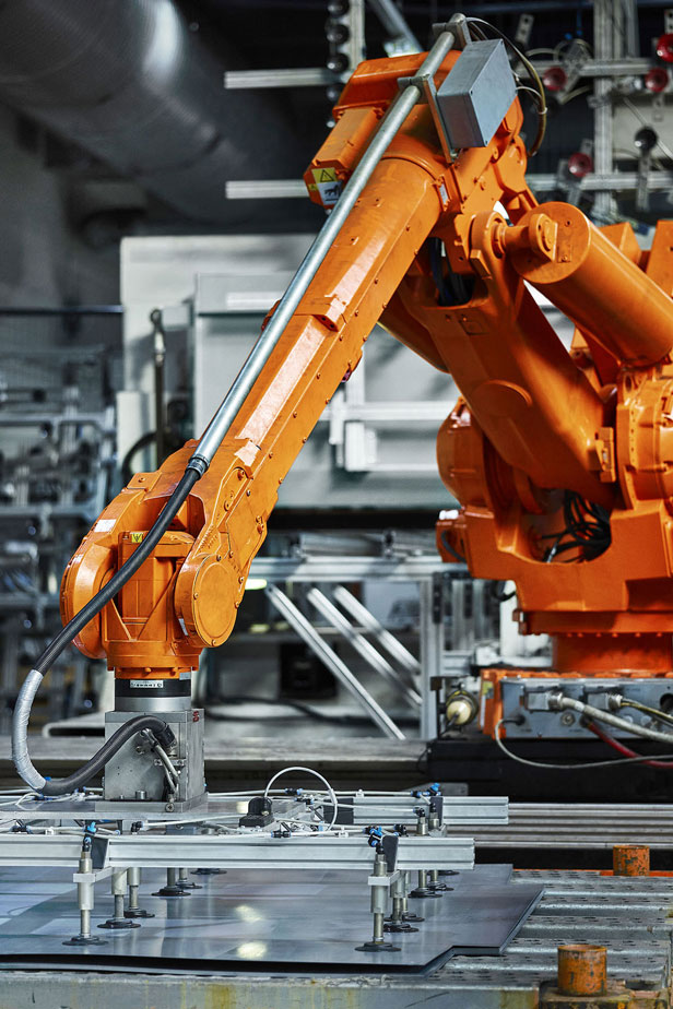 An orange industrial robotic arm operates over a workbench with clamps and fixtures in a factory setting; surrounded by machinery.