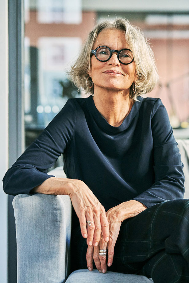 Woman sitting on a light gray sofa in a modern office setting; wearing a dark long-sleeve top and plaid pants.