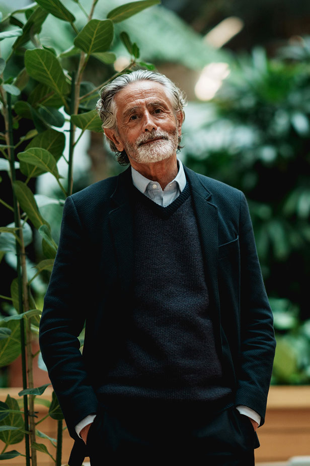Man wearing a dark suit and sweater standing in an indoor space filled with green plants