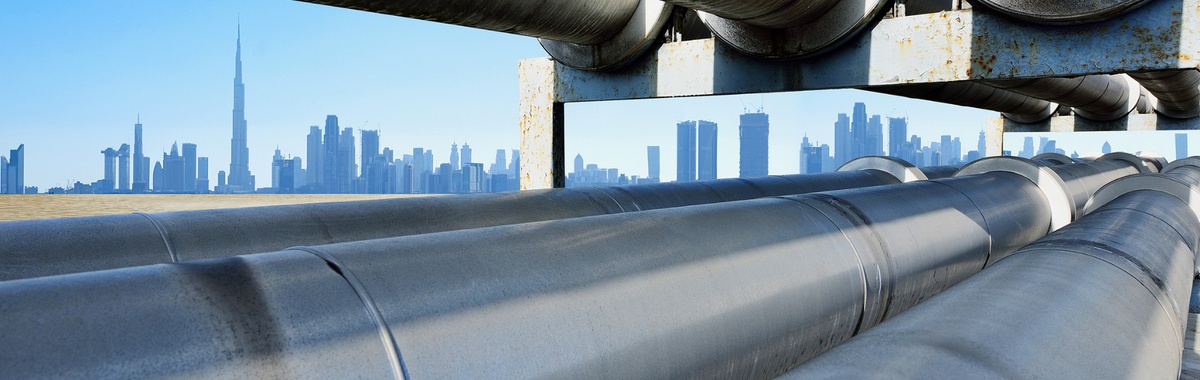 Large industrial pipelines extend toward a modern city skyline in the distance, with multiple steel pipes running across the foreground under a clear blue sky.