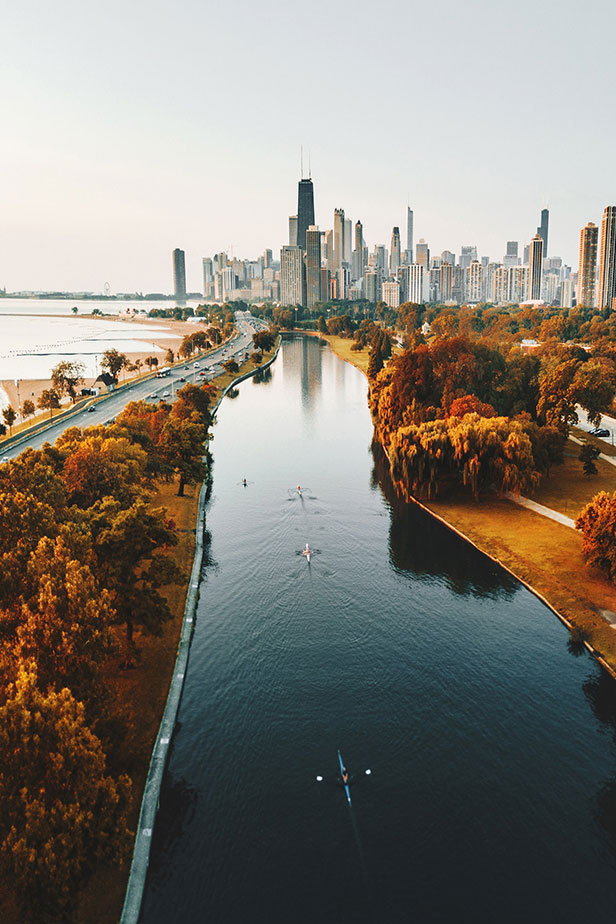 Highway with cars running alongside river and lake shoreline, with autumn foliage colours and skyline of Chicago in distance. 
