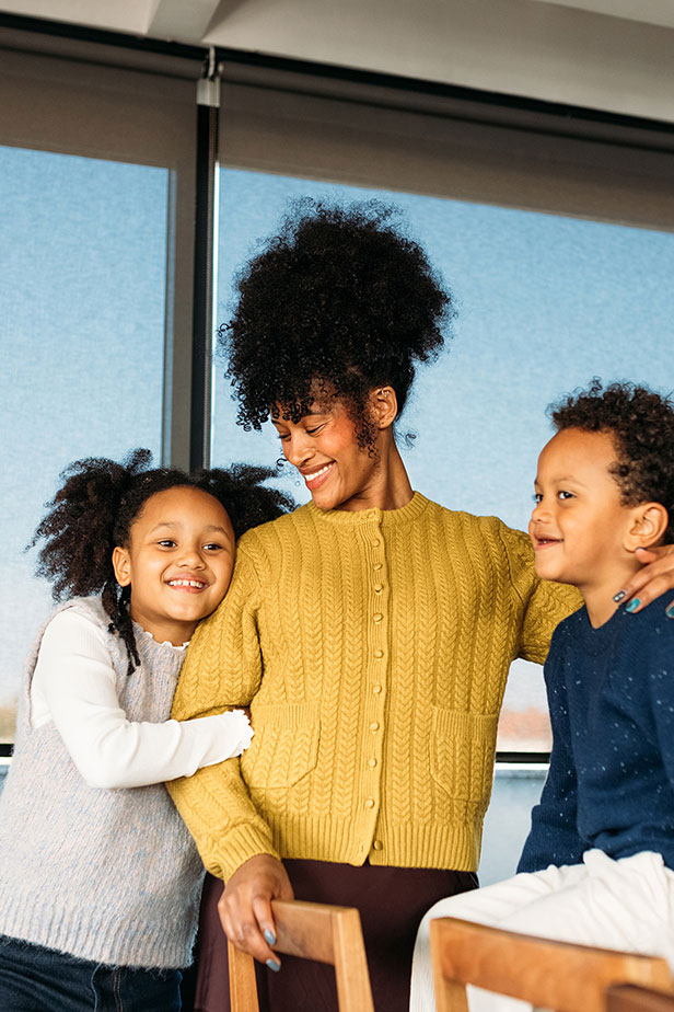 Woman standing indoors with two young children; one on each side; all dressed in casual clothing.