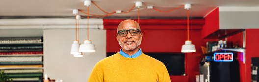 Smiling business owner in a yellow sweater standing inside his restaurant.