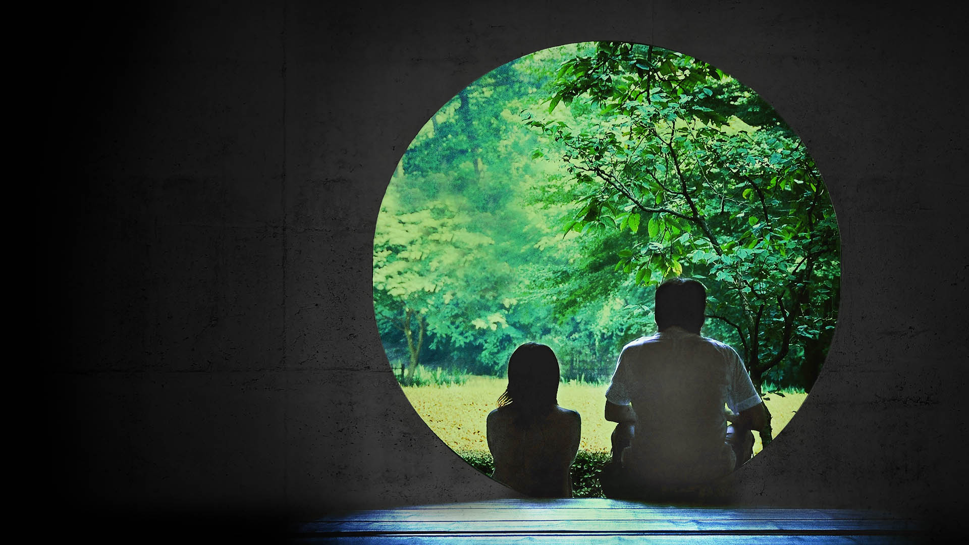 Two silhoetted people sit in front of a circular doorway, looking at green trees.