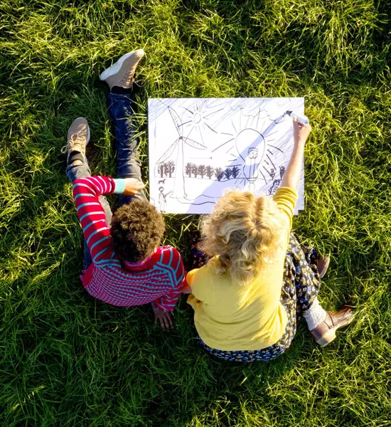 Zwei Kinder sitzen auf einer Wiese und haben ein selbstgezeichnetes Bild vor sich mit einer Windanlage