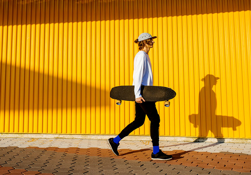 Po žltej stene kráča mladý muž so skateboardom pod pazuchou.