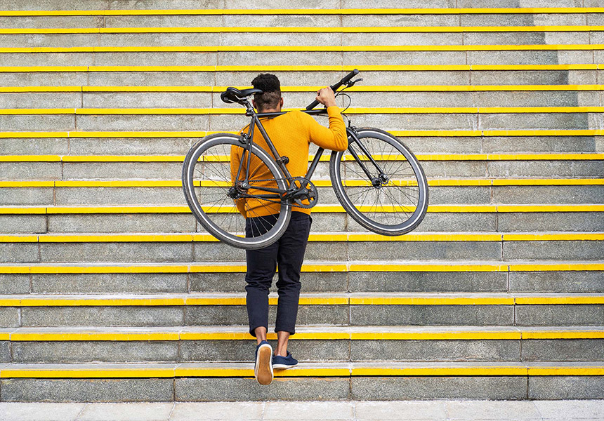 A man walks up a large outdoor staircase carrying his bicycle on his back.