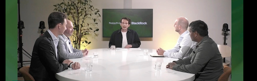 Vier mannen zitten rond een witte tafel in een vergaderruimte, terwijl één persoon in het midden spreekt. Op de achtergrond is een scherm te zien met de tekst 'Blockchain'.