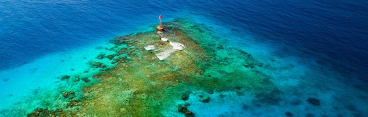 Luchtfoto van een koraalrif in helder turquoise water met een kleine rode boei.
