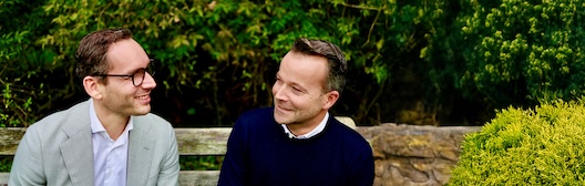 Twee mannen zitten buiten op een houten bank in een groene omgeving en lachen naar elkaar; op de achtergrond zijn een stenen muur en bomen te zien.