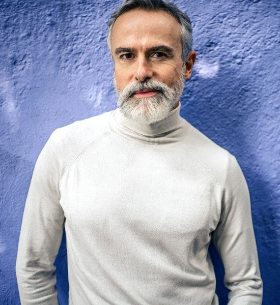 Ein Mann mit einem grauen Vollbart steht vor einer dunkelblauen Wand und blickt direkt in die Kamera.